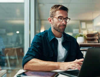 Mann mit Brille, dunkelblauem Hemd über weißem T-Shirt, sitzt am Schreibtisch und tippt auf Laptop; Tablet daneben.