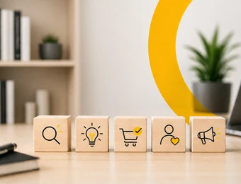 Fünf Bausteine mit Symbolen (Lupe, Glühbirne, Einkaufswagen, Profil, Megafon) auf Holztisch; gelber Bogen, Pflanze, Laptop