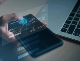 Eine Hand hält ein Smartphone mit digitalen Sicherheitsicons; Laptop im Hintergrund.