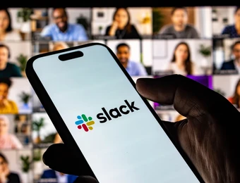 Nahaufnahme einer Hand, die ein Smartphone mit dem Slack-Logo hält; unscharfer Hintergrund mit vielen Videoteilnehmern.