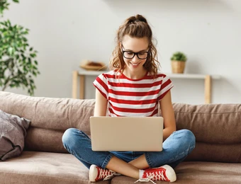 Person sitzt im Wohnzimmer auf einem Sofa, Laptop auf dem Schoß, rot-weiße Streifenbluse, Jeans, Brille, lächelnd.