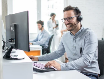 Mann mit Headset sitzt an Computerarbeitsplatz; im Hintergrund arbeiten weitere Personen im Büro.
