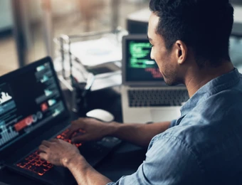Mann sitzt an zwei offenen Laptops und tippt auf der Tastatur; Codezeilen auf den Bildschirmen sichtbar.
