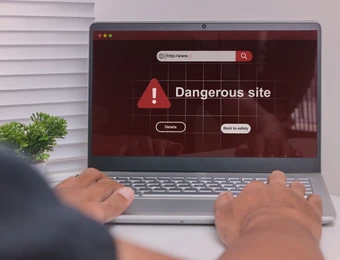 Laptop am Schreibtisch; Bildschirm zeigt rotes Warnfenster 'Dangerous site' mit Warnsymbol; Hände auf der Tastatur.
