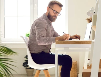Mann mit Bart und Brille sitzt an einem Schreibtisch, schreibt mit Stift, Laptop offen; heller Raum mit großen Fenstern.