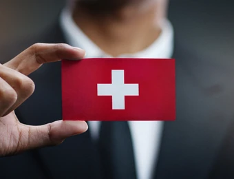 Eine Hand hält eine rote Karte mit weißem Kreuz vor unscharfem dunklem Hintergrund; Anzug sichtbar.