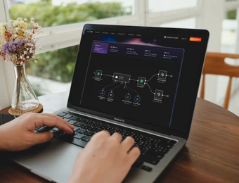 Person tippt auf MacBook Pro; Bildschirm zeigt dunkles Diagramm. Vase mit Blumen links, Fenster im Hintergrund.