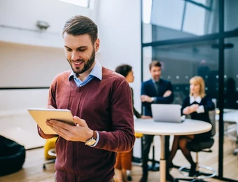 Mann in rotem Pullover hält Tablet; im Hintergrund stehen zwei Personen an einem runden Tisch im Büro.