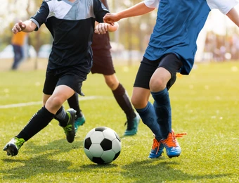 Fußballspieler rennen um den Ball auf grünem Rasen; Beine und Ball im Vordergrund.