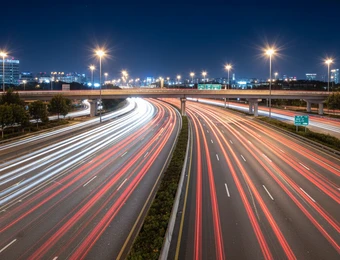 Nachtaufnahme einer mehrspurigen Autobahn mit roten und weißen Lichtstreifen, Brücke und Stadtlichter im Hintergrund.