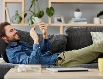 Mann liegt entspannt auf einem grauen Sofa und schaut auf sein Smartphone; im Hintergrund Regale mit Pflanzen.