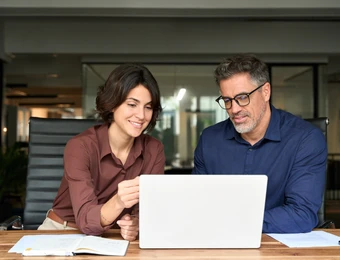 Eine Frau und ein Mann sitzen an einem Holztisch vor einem offenen Laptop; daneben liegen Unterlagen, Büro im Hintergrund.