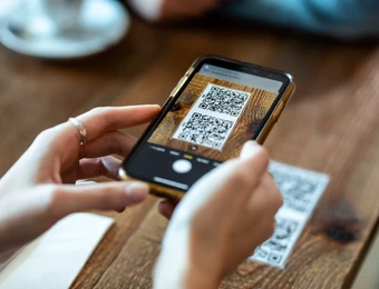 Nahaufnahme: Hände halten Smartphone, das einen QR-Code zeigt; weitere QR-Codes liegen auf einem Holztisch.