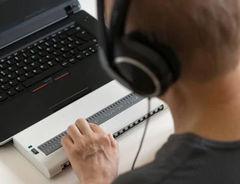 Person mit Kopfhörern vor Laptop; ein weißes Gerät mit vielen kleinen Tasten liegt auf dem Tisch.