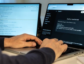 Person tippt am Laptop; zwei Monitore im Hintergrund, linker Bildschirm zeigt Code, rechter Bildschirm Chatfenster.