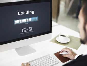Computerbildschirm mit Ladebildschirm und Fortschrittbalken (Loading, 70%); Maus, Tastatur, Tasse Kaffee, Person rechts.