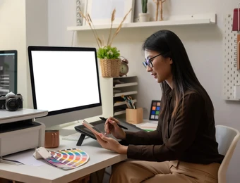 Eine Frau mit Brille sitzt am Schreibtisch und arbeitet an einem Grafiktablett; Monitor, Farbmuster, Kamera und Drucker.