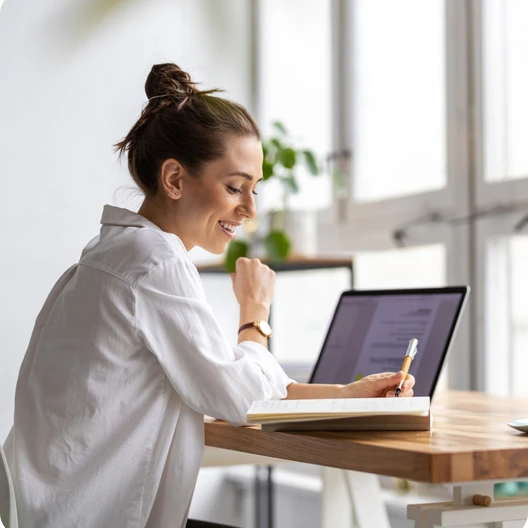 Eine Frau sitzt an einem Holztisch, schreibt in ein Notizbuch; Laptop geöffnet, helles Büro mit Fenster.
