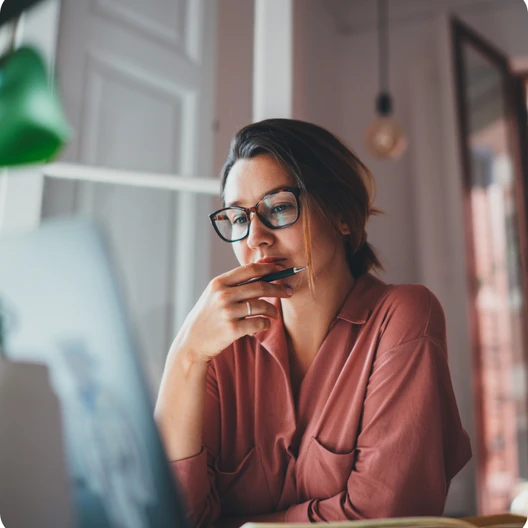 Frau mit Brille in rosa Oberteil sitzt an einem Tisch vor Laptop, rechte Hand am Kinn, Blick nach unten.