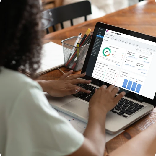 Person tippt an einem Laptop; Bildschirm zeigt Diagramme und Dashboards, Tisch mit Stiftehalter und Notizbuch.