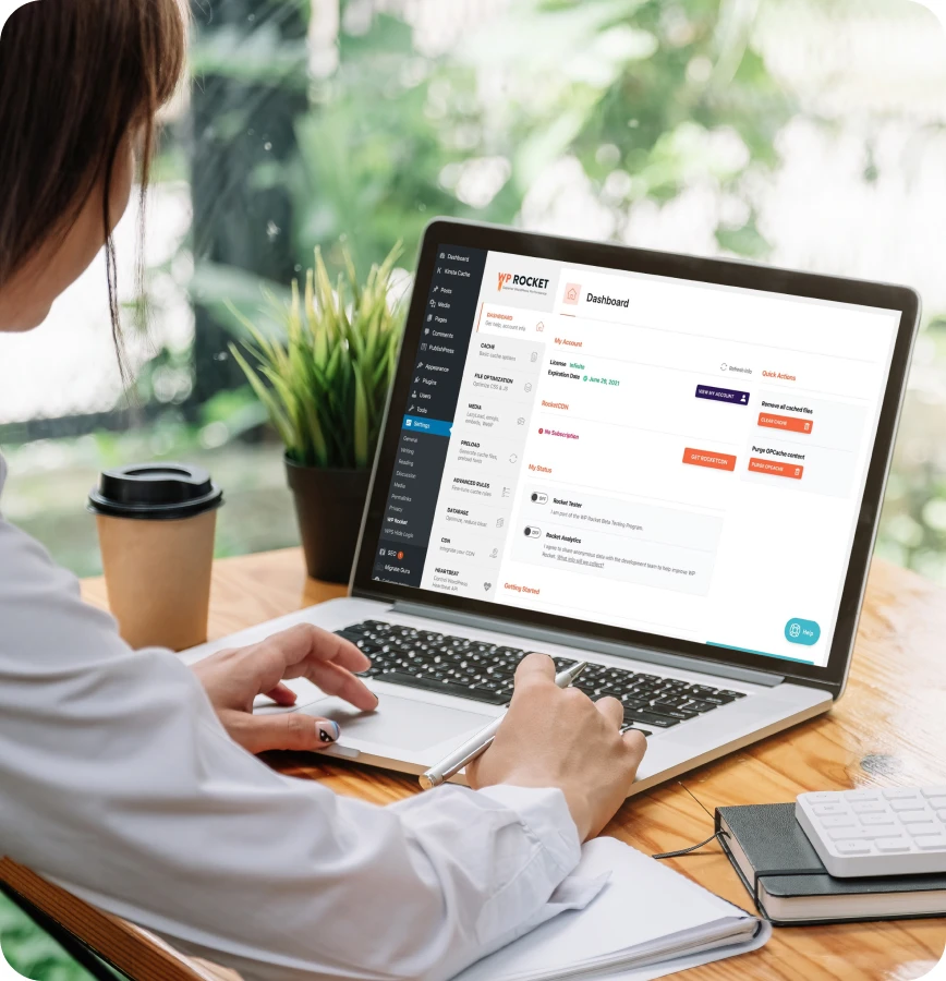 Person von hinten sitzt an Holztisch und nutzt Laptop mit Dashboard; Kaffeebecher, Pflanze und Notizbuch daneben.