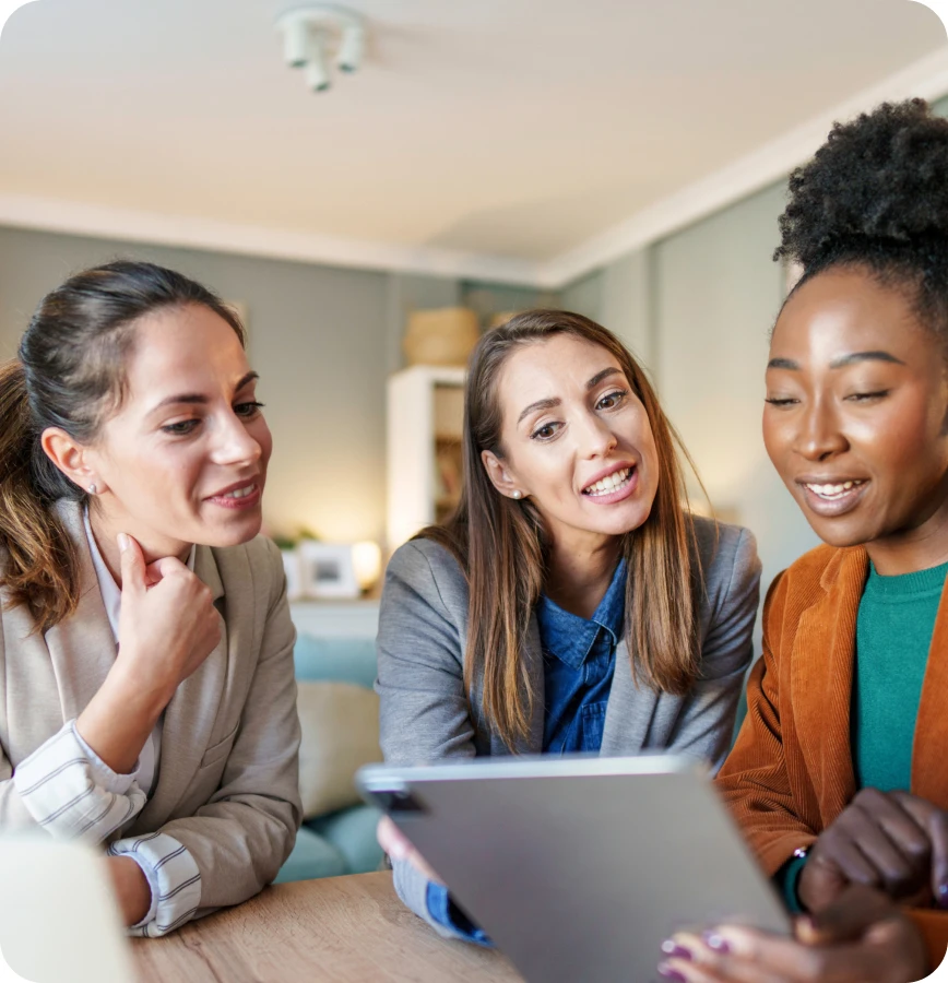 Drei Frauen sitzen an einem Tisch und schauen auf ein Tablet; heller Innenraum im Hintergrund.