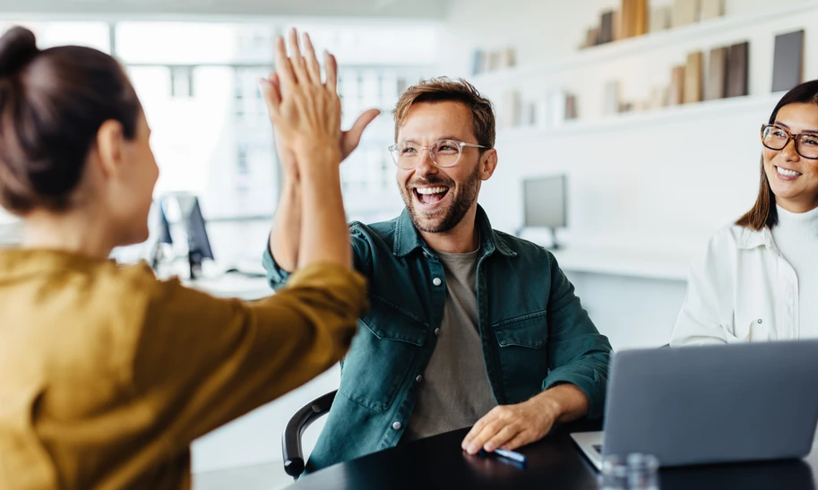 Drei Personen in einem Büro; Mann und Frau geben sich High-Five über dem Tisch, daneben lächelt eine Frau; Laptop sichtbar.