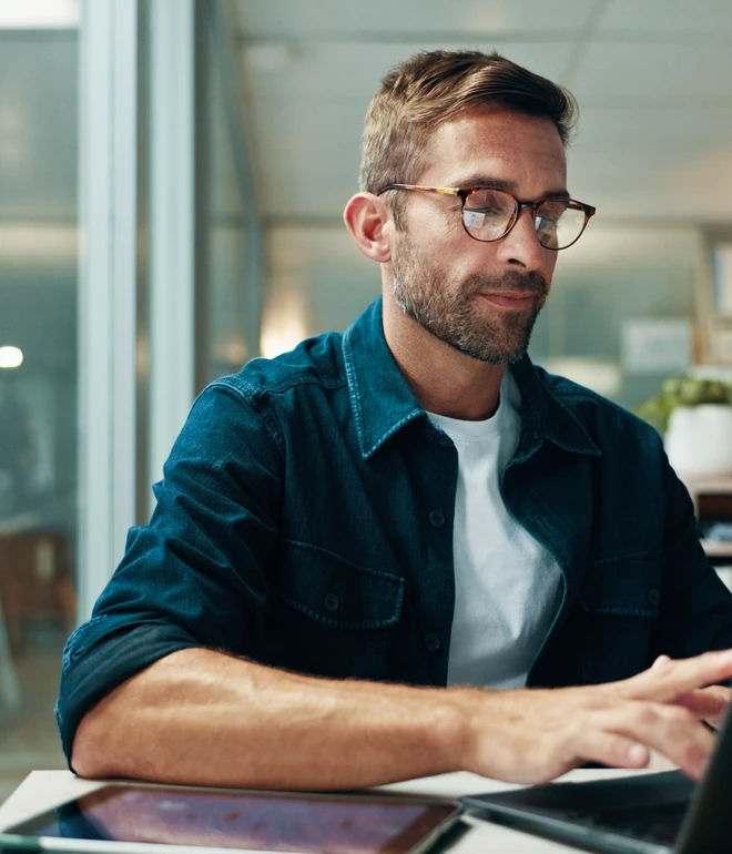 Mann mit Brille, dunkelblauem Hemd über weißem T-Shirt, sitzt am Schreibtisch und tippt auf Laptop; Tablet daneben.