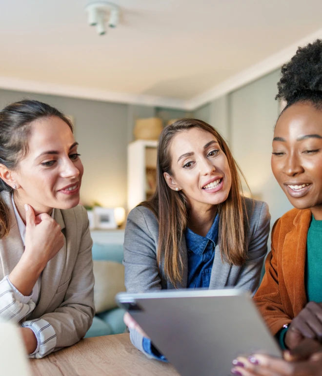 Drei Frauen sitzen an einem Tisch und schauen auf ein Tablet; heller Innenraum im Hintergrund.