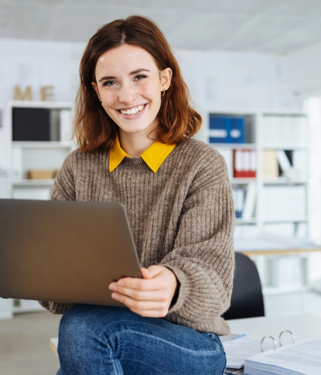 Frau mit braunem Pullover und gelbem Kragen hält Laptop in hellem Büro; Regale im Hintergrund.
