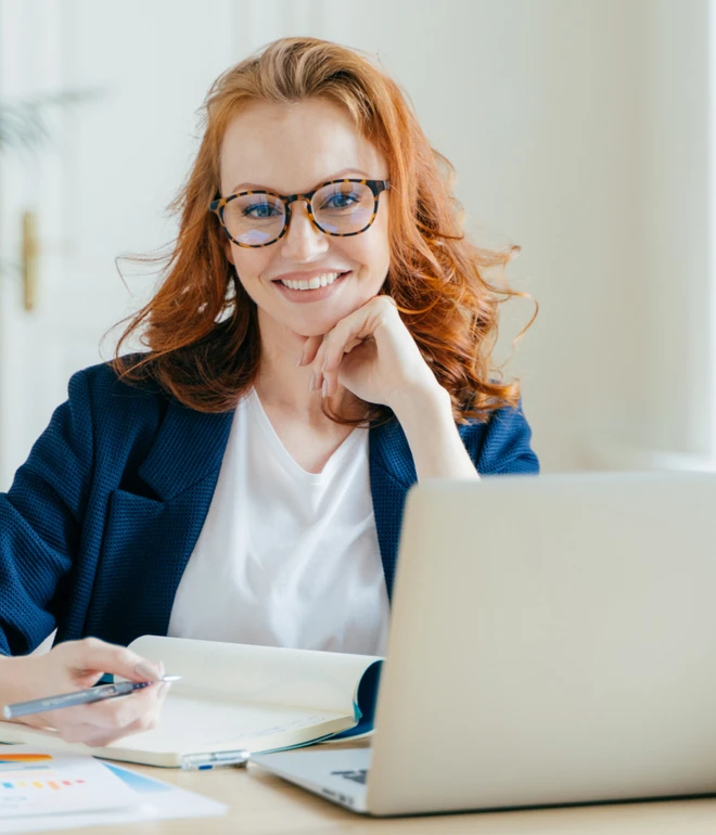 Frau mit roten Haaren und Brille, blauem Blazer, weißem Top, sitzt am Schreibtisch mit Laptop und Notizblock, lächelt.