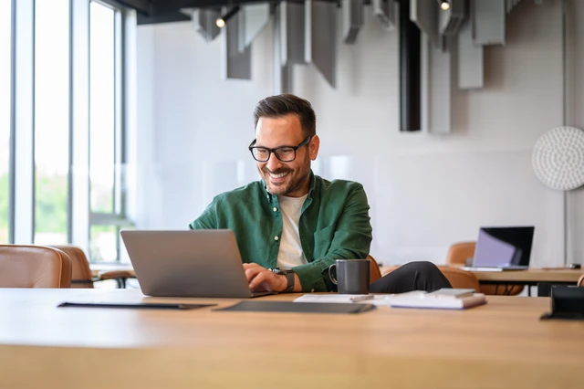 Mann mit Brille sitzt an einem Laptop am Tisch in einem hellen Büro; grüne Jacke, Kaffeebecher im Vordergrund.