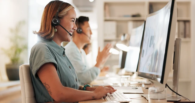 Eine Frau im Vordergrund mit Headset am Computer; helles Büro mit weiteren Mitarbeitenden und Monitoren.