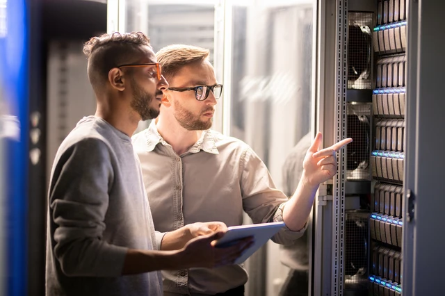 Zwei Männer vor einem Serverrack in einem Rechenzentrum; einer hält ein Tablet und zeigt auf die Server.