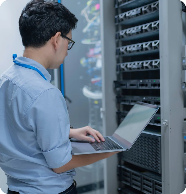 Person mit Brille und blauem Hemd arbeitet mit Laptop vor Serverracks.
