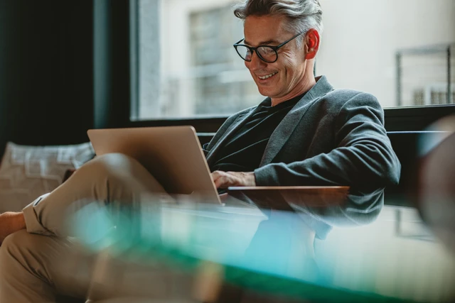 Mann mit Brille und grauen Haaren sitzt mit Laptop auf dem Schoß und lächelt; Fenster im Hintergrund.