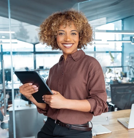 Fröhliche Frau mit Locken hält Tablet, steht neben Schreibtisch mit Laptop in modernem Büro.