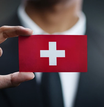 Eine Hand hält eine rote Karte mit weißem Kreuz vor unscharfem dunklem Hintergrund; Anzug sichtbar.