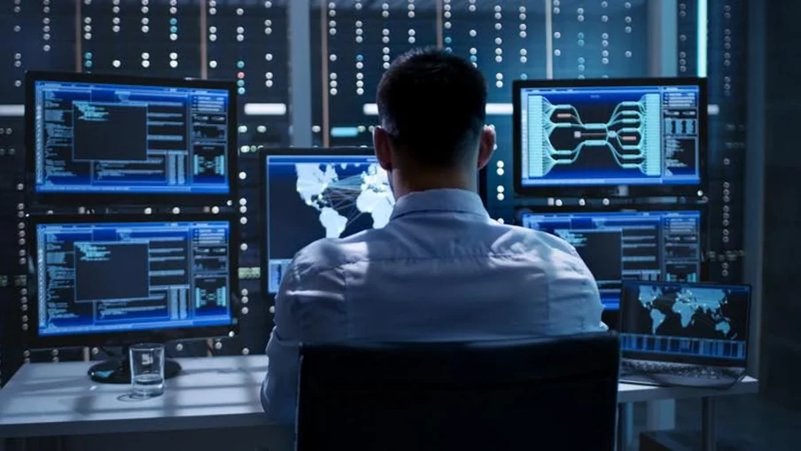 Person von hinten vor mehreren Monitoren mit Code, Weltkarte und Diagrammen in dunklem Serverraum.