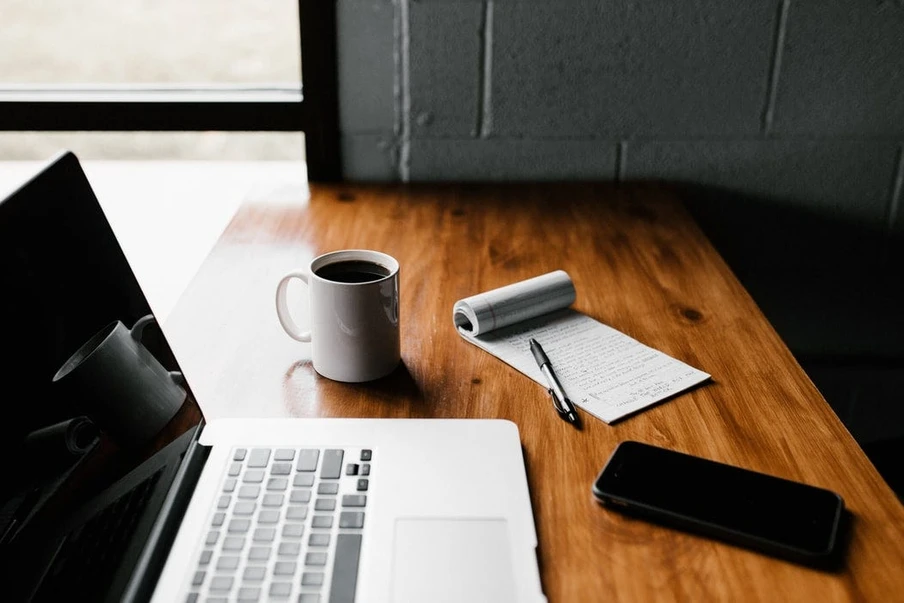 Laptop auf Holztisch, Kaffeebecher, Notizblock mit Stift, Papierrolle und Smartphone; Fenster im Hintergrund.