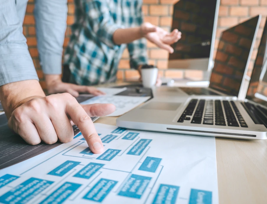 Nahaufnahme: Eine Hand zeigt auf blaue Karten auf Papier auf einem Tisch; Laptop und Kaffeetasse im Hintergrund.