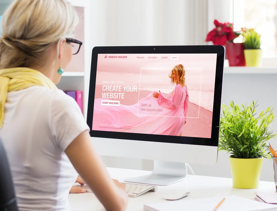Person von hinten mit hellem Haar und Brille vor einem iMac; Bildschirm zeigt pinke Website, Schreibtisch mit Pflanze.