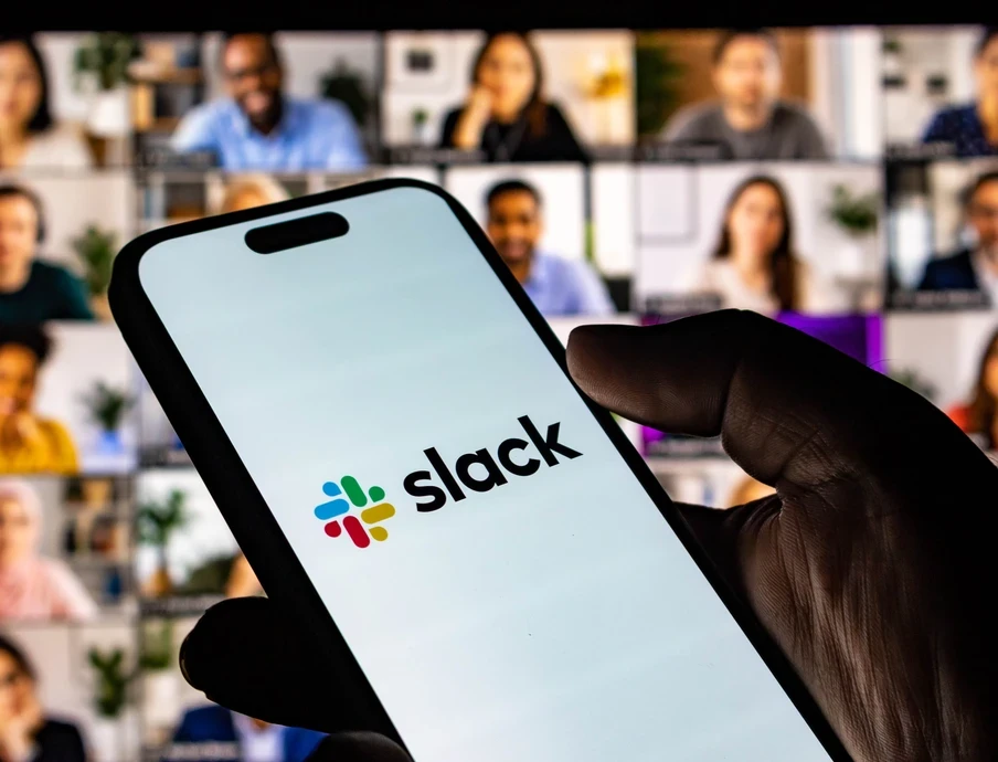 Nahaufnahme einer Hand, die ein Smartphone mit dem Slack-Logo hält; unscharfer Hintergrund mit vielen Videoteilnehmern.