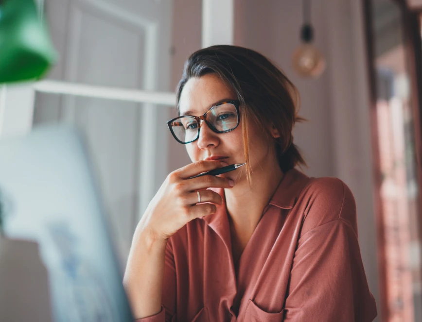 Frau mit Brille in rosa Oberteil sitzt an einem Tisch vor Laptop, rechte Hand am Kinn, Blick nach unten.