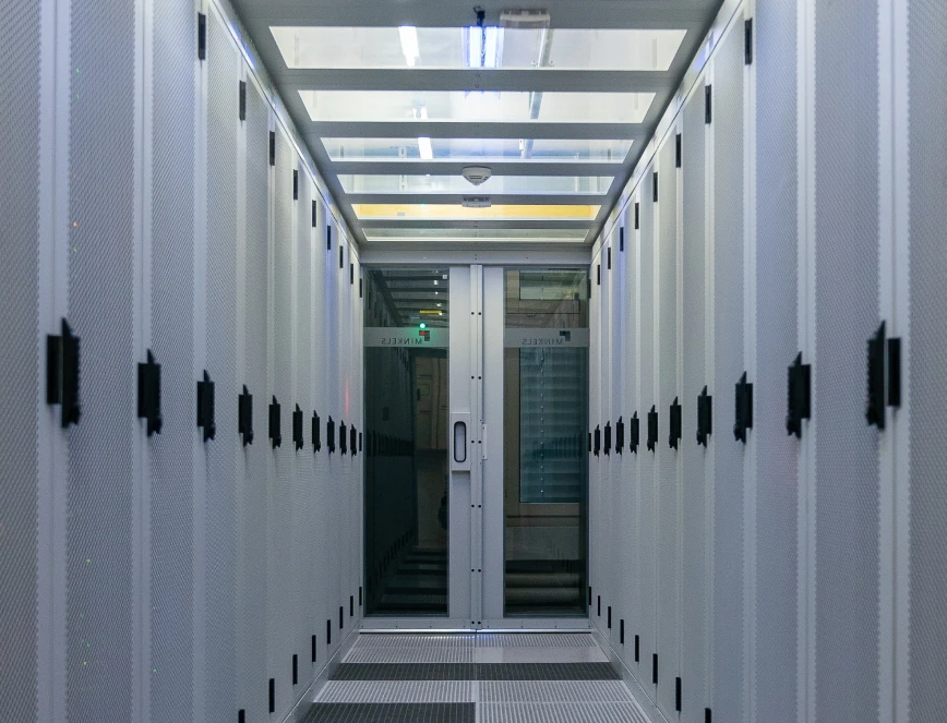 Gangkorridor mit Serverracks auf beiden Seiten, metallene Gehäuse, hell beleuchtet, Tür am Ende.