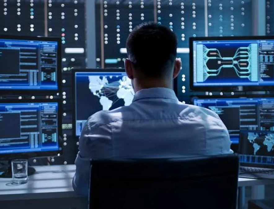Person von hinten vor mehreren Monitoren mit Code, Weltkarte und Diagrammen in dunklem Serverraum.