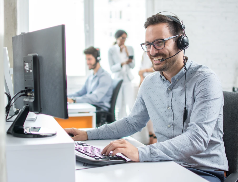 Mann mit Headset sitzt an Computerarbeitsplatz; im Hintergrund arbeiten weitere Personen im Büro.