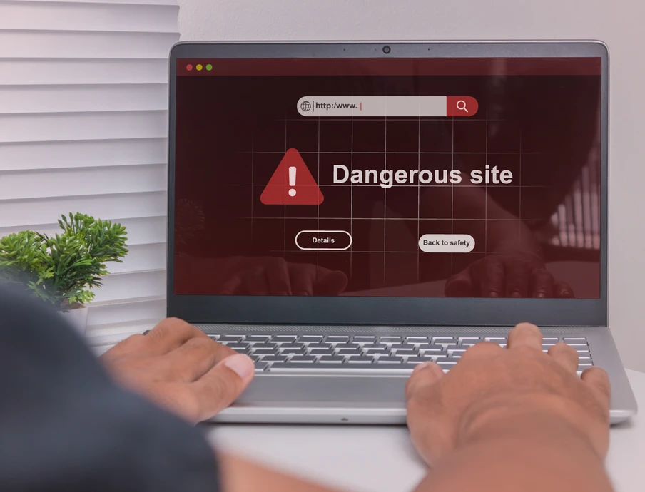 Laptop am Schreibtisch; Bildschirm zeigt rotes Warnfenster 'Dangerous site' mit Warnsymbol; Hände auf der Tastatur.