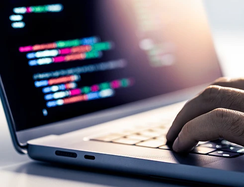 Nahaufnahme eines Laptops; Bildschirm zeigt farbigen Code, eine Hand tippt auf der Tastatur.