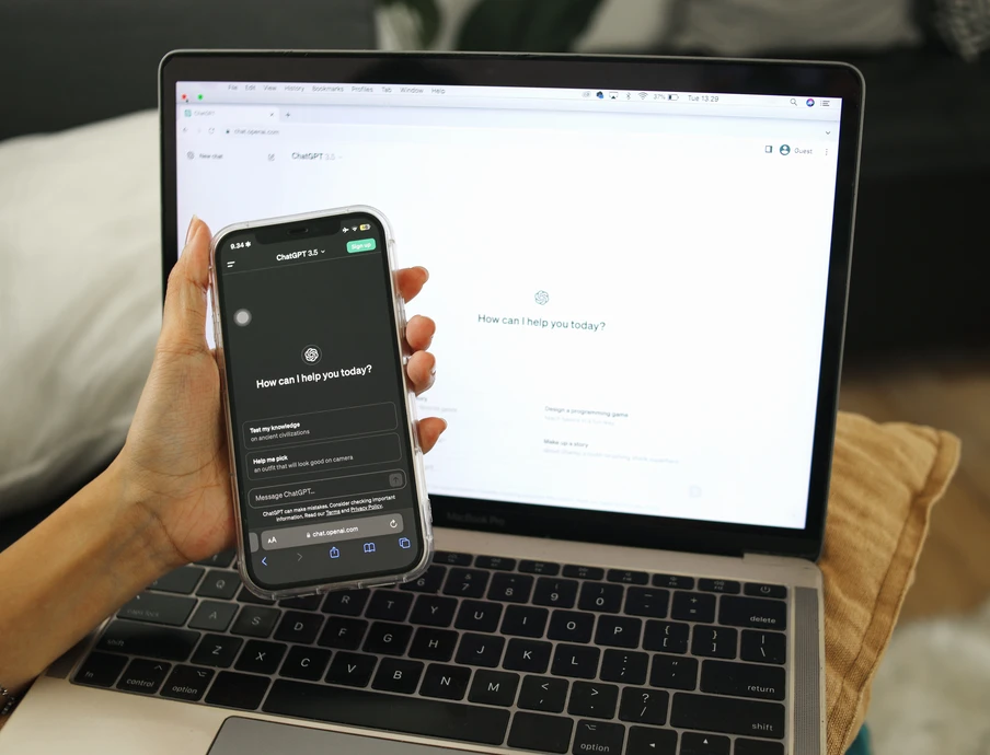 Eine Hand hält ein Smartphone vor einem geöffneten Laptop; Smartphone-Bildschirm zeigt Chat-Nachricht 'How can I help you today?'